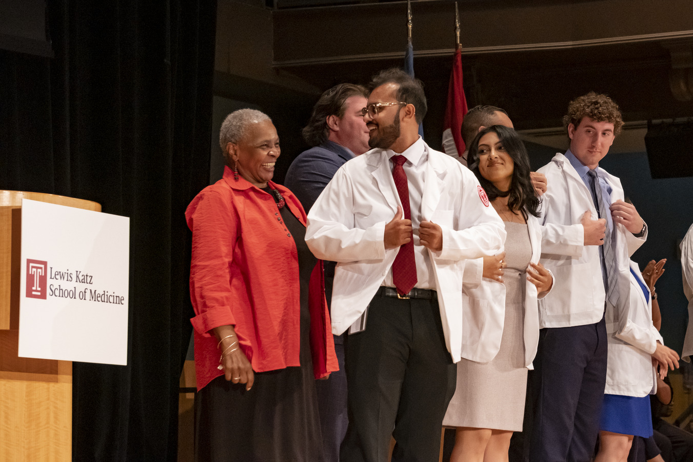 A smiling student being coated by Dr. Stewart in a group of students receiving white coats from other people