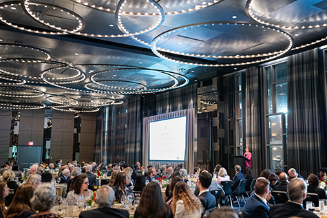 a woman giving a speech at a podium in front of the dinner guests in a grant room with ornate lighting on the ceiling