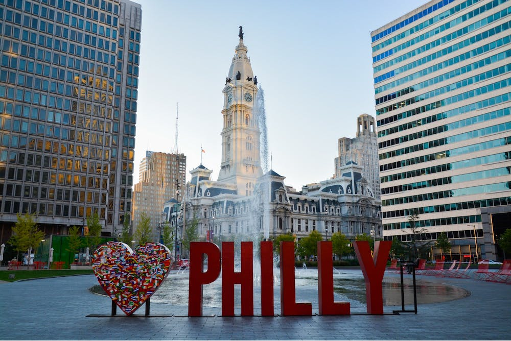 Red Philly sign with a heart next to and tall modern buildings on each side and a clock tower in the background.