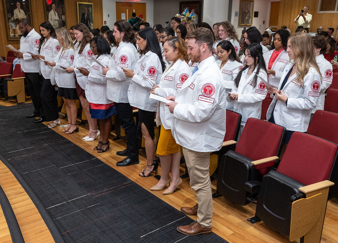 Students wearing white coats standing over their chairs reading a letter of paper on their hands